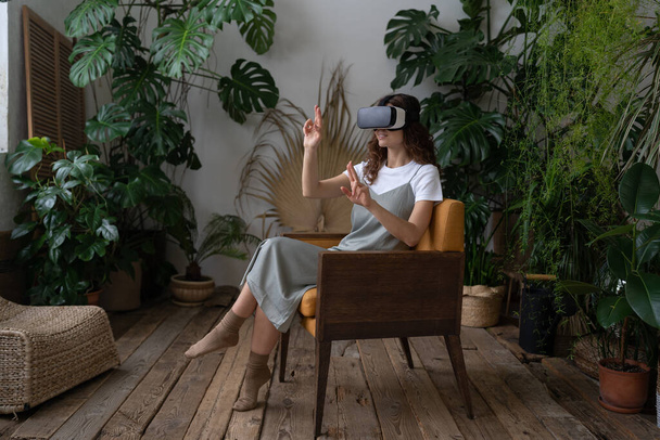 VR and AR technologies. Young impressed woman in virtual reality glasses touching air, sitting in armchair in cozy indoor garden with abundance of tropical greenery, playing game or watching video - Photo, Image