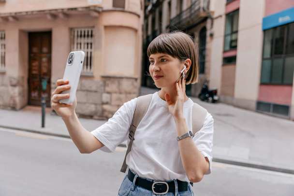 Jeune fille excitée en t-shirt blanc dans les écouteurs et t-shirt blanc tient smartphone. Femme joyeuse en tenue hipster élégant sourit à l'extérieur.  - Photo, image