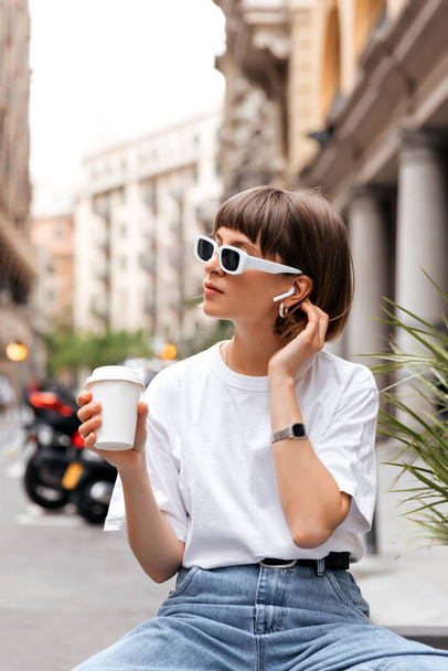 Cute dark-haired girl in sunglasses drinking coffee in good sunny day. Portrait of jocund female model in stylish outfit holding coffee. - Photo, Image