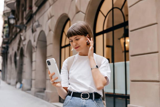 Beautiful young caucasian woman walks near hotel building. Portrait of successful stylish woman holding smartphone and touching headphones in her hand on way to work on city street. - Photo, Image