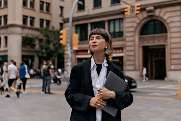 Obchodní žena v černé bundě a bílé košili s rovnými krátkými vlasy drží notebook procházky v obchodní čtvrti. Úspěšná pozitivní dívka jít po práci venku. - Fotografie, Obrázek