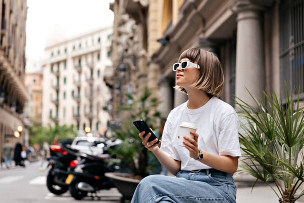 Jeune femme caucasienne passer du temps libre dans la ville européenne en été jour téléphone portable tout en étant assis à l'extérieur. Souriante fille se détend à l'extérieur en buvant du café. Enseignement à distance en ligne. - Photo, image