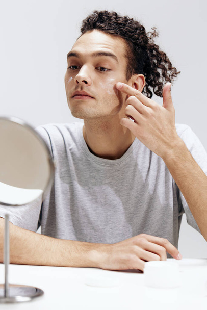chlap s kudrnatými vlasy nanášení krém na obličej a při pohledu do zrcadla kosmetologie izolované pozadí. Kvalitní fotografie - Fotografie, Obrázek