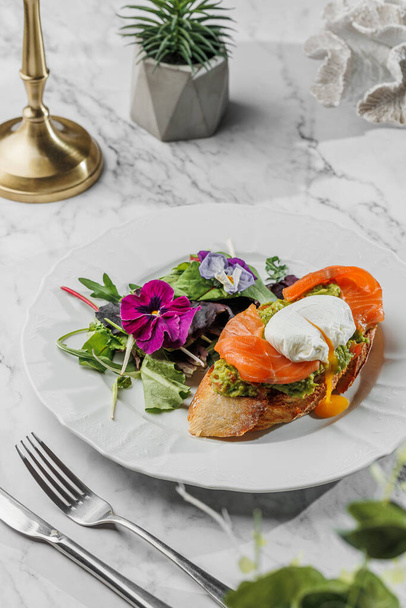 Bruschetta with Poached egg and fresh salmon , vegetarian salad with greens at the white plate on light marble background. Healthy sea food, hard light, restaurant decor - Фото, зображення