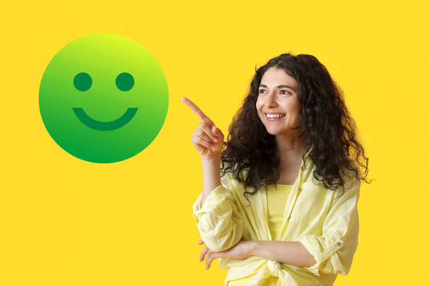 Beautiful young woman pointing at smiling emoticon on yellow background - Photo, Image