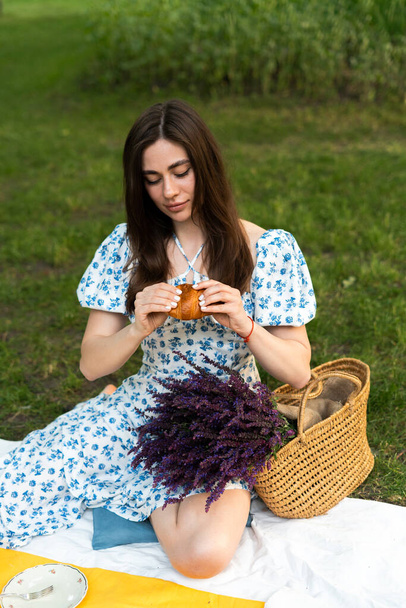 een jong meisje in een blauwe jurk breekt een croissant bij een picknick in de natuur - Foto, afbeelding