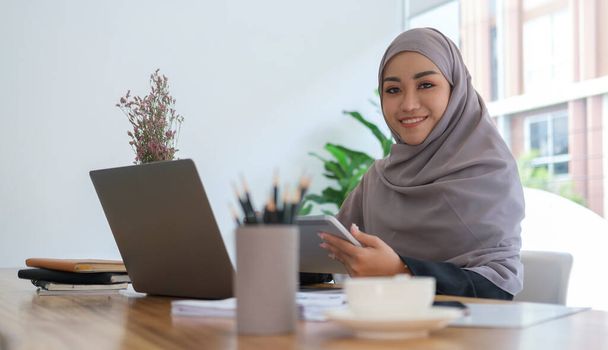 Portret van lachende jonge islamitische zakenvrouw in hijab zittend aan kantoor bureau met computer laptop en glimlachend op camera. - Foto, afbeelding