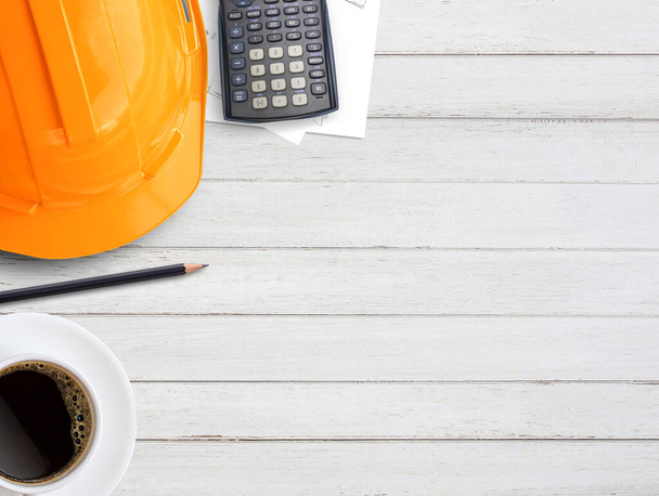 Pomarańczowy kask bezpieczeństwa lub twardy kapelusz, kawa, kalkulator i plany papieru budowlanego na tle drewnianego stołu. Pomysł na budowanie biznesu, koncepcja budownictwa. Widok z góry z miejsca do kopiowania do użytku. - Zdjęcie, obraz