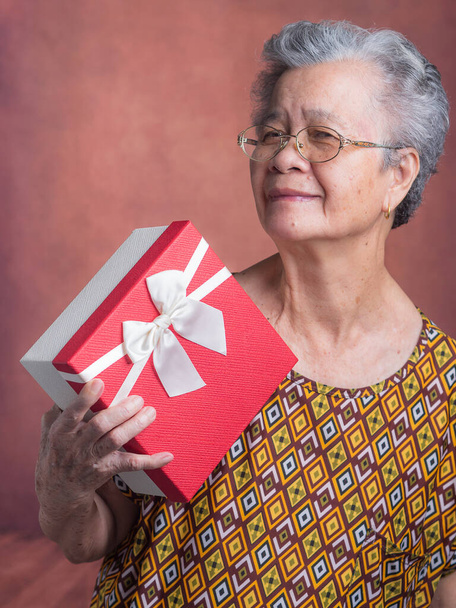 Portret starszej Azjatki trzymającej czerwone pudełko z prezentami i patrzącej w kamerę stojącej w studiu. Pojęcie festiwalu, rocznicy i osób w podeszłym wieku. - Zdjęcie, obraz