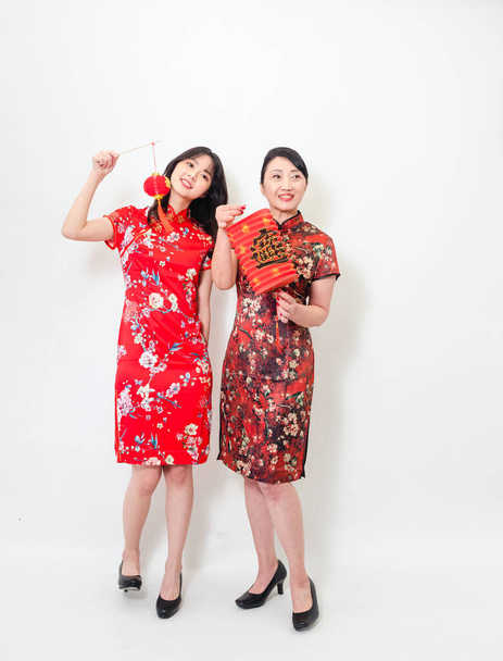 Ritratto di giovane donna asiatica e donna anziana che indossano entrambi l'abito tradizionale cheongsam qipao tenere lanterna di carta rossa con parola che significa buona fortuna e tesoro su sfondo bianco. - Foto, immagini