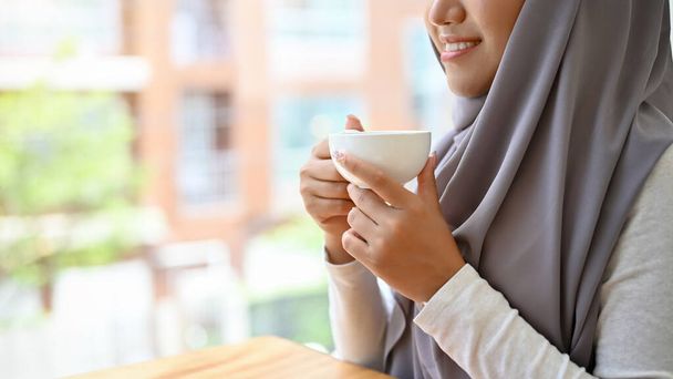 Beautiful attractive muslim woman with hijab relaxes sipping a morning coffee at home or office. cropped image, hands holding a coffee cup. - Foto, imagen