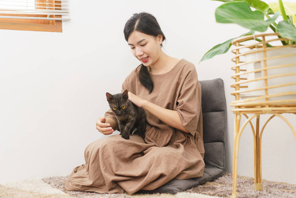 Молода азіатка сидить на дивані на підлозі і за допомогою руки, щоб погладити кішку.. - Фото, зображення