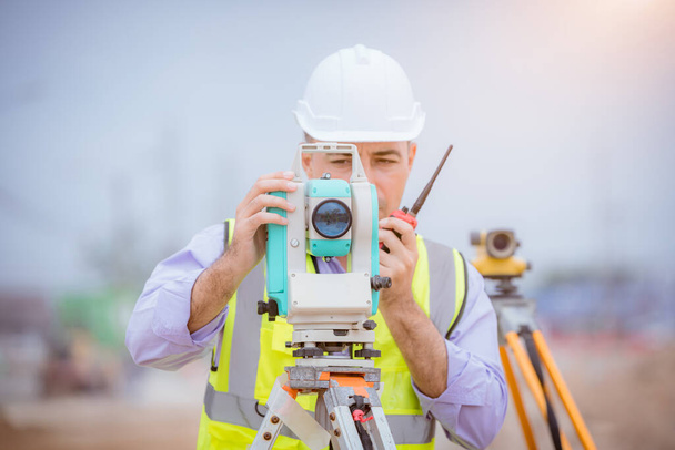 Surveyor engineers wearing safety uniform ,helmet and radio communication with equipment theodolite to measurement positioning on the construction site of the road with construct machinery background. - Фото, изображение