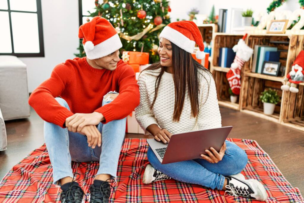 Young latin couple smiling happy and using laptop sitting by christmas tree at home. - Photo, image