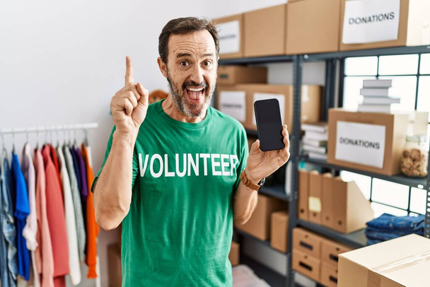 Hombre de mediana edad con barba con camiseta voluntaria sosteniendo el teléfono inteligente apuntando con el dedo hacia arriba con una idea exitosa. exitado y feliz. número uno.  - Foto, Imagen
