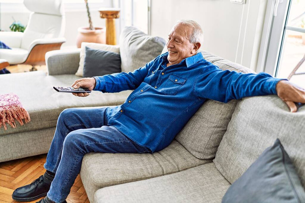 Hombre mayor con el pelo gris sentado en el sofá en la sala de estar de su casa. Hombre maduro sonriendo feliz en casa. - Foto, Imagen