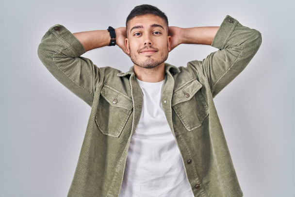 Jeune homme hispanique debout sur fond isolé relaxant et étirant, bras et mains derrière la tête et le cou souriant heureux  - Photo, image