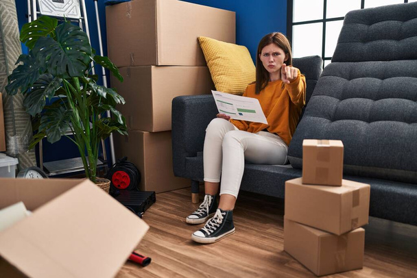 Young brunette woman sitting on the sofa at new home looking at documents pointing with finger to the camera and to you, confident gesture looking serious  - Foto, afbeelding