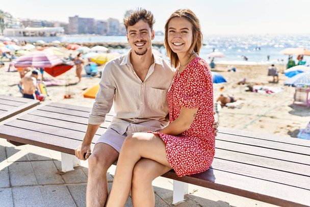 Νεαρό ζευγάρι ισπανόφωνων σε διακοπές χαμογελώντας χαρούμενο να κάθεται στον πάγκο στην παραλία - Φωτογραφία, εικόνα