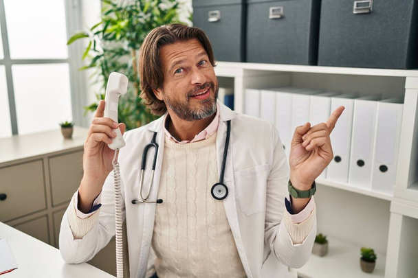 Bell'uomo medico di mezza età che parla al telefono alla clinica sorridendo felice indicando con mano e dito di lato  - Foto, immagini