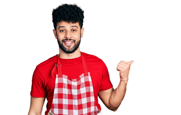 młody arabski człowiek z brodą ubrany w fartuch kucharski uśmiechnięty ze szczęśliwą twarzą patrzący i wskazujący na bok z kciukiem w górze.  - Zdjęcie, obraz