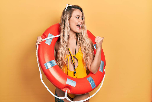 Beautiful young blonde woman wearing bikini and holding lifeguard float pointing thumb up to the side smiling happy with open mouth  - Valokuva, kuva
