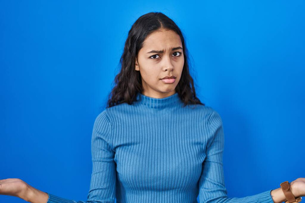 Молодая бразильская женщина стоит на голубом изолированном фоне, ничего не зная и путаясь с открытыми объятиями, понятия не имею.  - Фото, изображение
