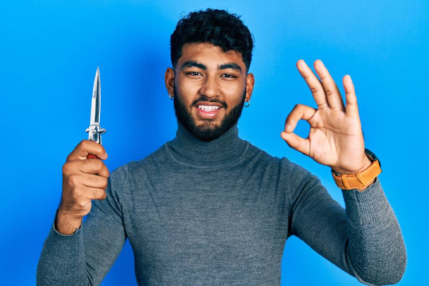 Άραβας με γενειάδα κρατώντας μαχαίρι τσέπης κάνει ok υπογράψει με τα δάχτυλα, χαμογελώντας φιλικό gesturing εξαιρετικό σύμβολο  - Φωτογραφία, εικόνα