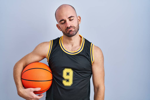 Молодой лысый мужчина с бородой в баскетбольной форме держит мяч, выглядящий сонным и уставшим, уставшим от усталости и похмелья, ленивыми глазами по утрам.  - Фото, изображение
