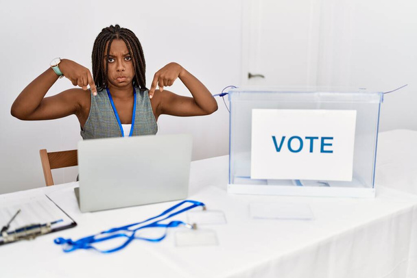 Mujer afroamericana joven trabajando en elecciones políticas sentada por votación apuntando hacia abajo mirando triste y molesto, indicando dirección con los dedos, infeliz y deprimido.  - Foto, imagen