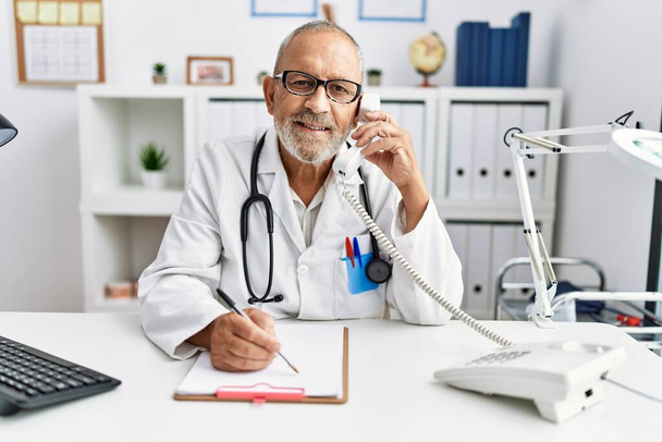 Ένας ηλικιωμένος γκριζομάλλης με στολή γιατρού που μιλάει στο τηλέφωνο στην κλινική. - Φωτογραφία, εικόνα