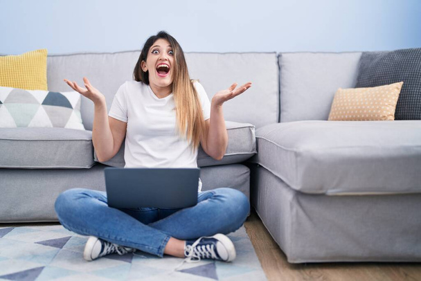 Young brunette woman sitting on the floor at home using laptop celebrating victory with happy smile and winner expression with raised hands  - Zdjęcie, obraz
