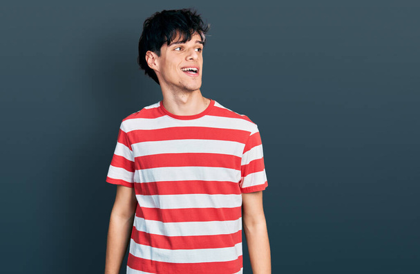 Handsome hipster young man wearing casual striped t shirt looking away to side with smile on face, natural expression. laughing confident.  - Zdjęcie, obraz