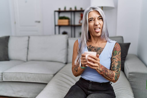 Mujer de pelo gris de mediana edad bebiendo café sentada en el sofá en casa sacando la lengua feliz con expresión divertida. concepto de emoción.  - Foto, Imagen