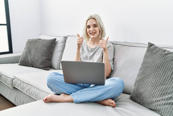 Молода біла жінка використовує ноутбук вдома, сидячи на дивані знак успіху робить позитивний жест рукою, великі пальці вгору посміхаються і щасливі. веселий вираз і жест переможця
.  - Фото, зображення