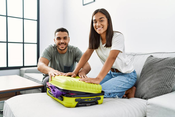 Latino hombre y mujer pareja sonriendo confiado cerca viaje maleta en casa - Foto, imagen