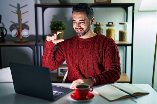 Young hispanic man with beard using computer laptop at night at home smiling and confident gesturing with hand doing small size sign with fingers looking and the camera. measure concept.  - Zdjęcie, obraz