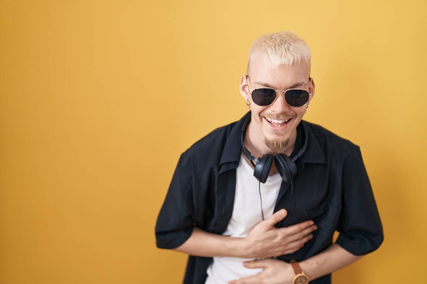 Young caucasian man wearing sunglasses standing over yellow background smiling and laughing hard out loud because funny crazy joke with hands on body.  - Fotó, kép