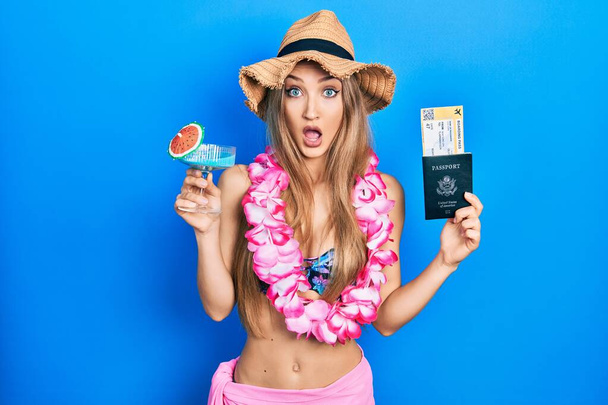 Chica rubia joven con bikini sosteniendo cóctel y pasaporte asustado y sorprendido con la expresión sorpresa y asombrado, miedo y cara emocionada.  - Foto, imagen