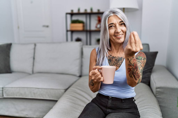 Середнього віку сіроока жінка п'є каву, сидячи на дивані вдома роблячи італійський жест з рукою і пальцями впевнений вираз
  - Фото, зображення
