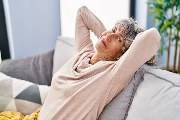 Orta yaşlı bir kadın, elleri başının üstünde, evinde kanepede oturuyor. - Fotoğraf, Görsel