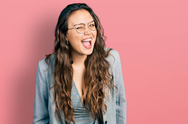 Молодая латиноамериканка в деловой одежде и очках подмигивает, глядя в камеру с сексуальным выражением лица, веселым и счастливым лицом.  - Фото, изображение