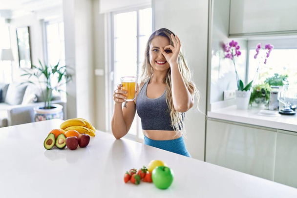 Young caucasian fitness woman wearing sportswear drinking healthy orange juice smiling happy doing ok sign with hand on eye looking through fingers  - Фото, изображение