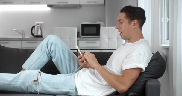 Happy young man receiving good news. Excited overjoyed male winner celebrating success looking at smartphone and making yes gesture while resting at home on couch. Mobile victory concept. 4K - Footage, Video