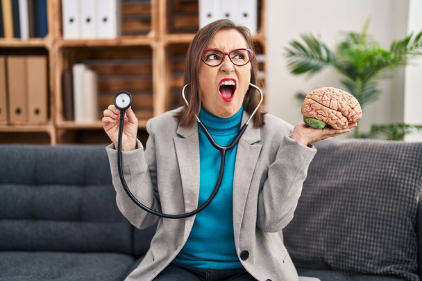 Middle age hispanic woman working at therapy office holding brain angry and mad screaming frustrated and furious, shouting with anger. rage and aggressive concept.  - Zdjęcie, obraz