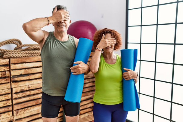Middle age couple holding yoga mat smiling and laughing with hand on face covering eyes for surprise. blind concept.  - Foto, afbeelding