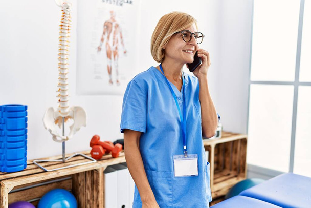 Keski-ikä blondi nainen yllään fysioterapeutti univormu puhuu älypuhelimeen klinikalla - Valokuva, kuva