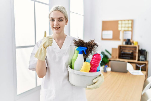 Young caucasian woman wearing cleaner uniform holding cleaning products cleaning office smiling happy and positive, thumb up doing excellent and approval sign  - Photo, Image