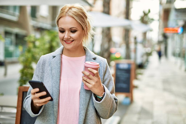 Young blonde businesswoman using smartphone drinking coffee at the city. - 写真・画像