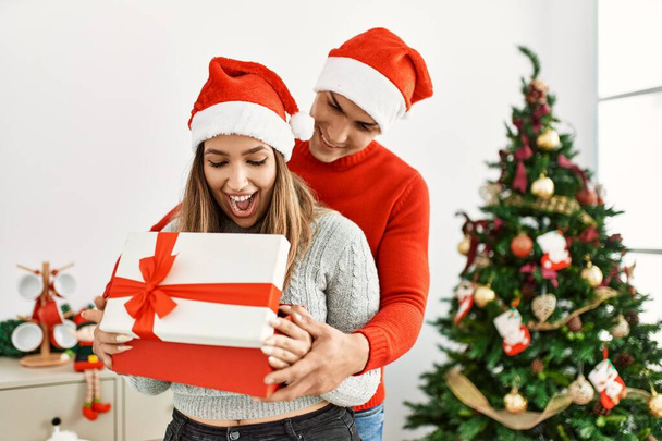 Νεαρό ζευγάρι αγκαλιάζει και χαμογελά ευτυχισμένο. Φορώντας χριστουγεννιάτικο καπέλο κρατώντας δώρο στο σπίτι. - Φωτογραφία, εικόνα